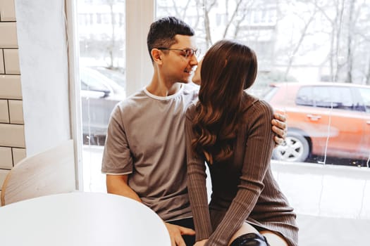 A couple shares a warm moment in a cafe, embracing and smiling at each other. Outside, a wintry scene unfolds with cars parked and snow falling gently.