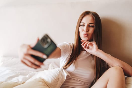 A young woman is playfully taking a selfie in a comfortable bedroom. She is sitting on the bed with white sheets, dressed casually, and smiling as she poses with her phone.