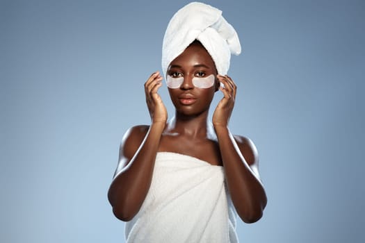 A woman stands confidently with a towel wrapped on her head and shoulders, applying a skincare mask while enjoying a self-care moment in a bright area.