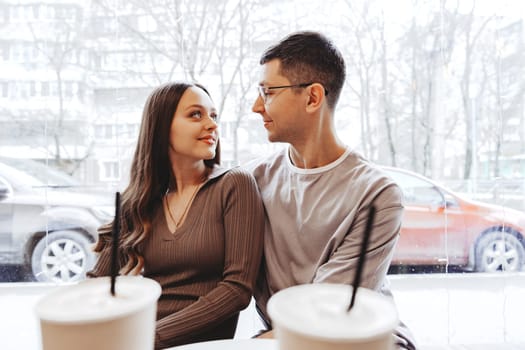 Two individuals share a warm moment in a cafe, gazing into each other's eyes over coffee. Outside, snowflakes fall softly, creating a picturesque winter backdrop.