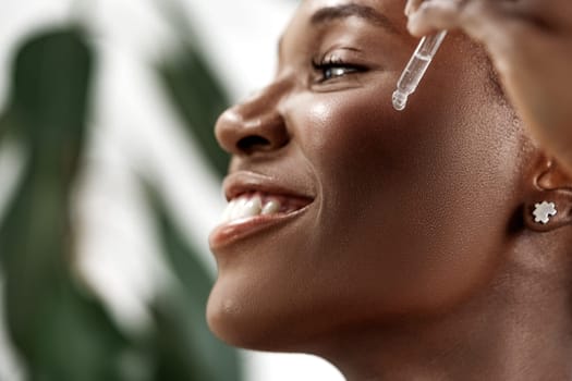 A woman with a joyful expression is applying a dropper serum to her face, surrounded by greenery. Sunlight highlights her radiant skin as she enjoys her skincare routine.