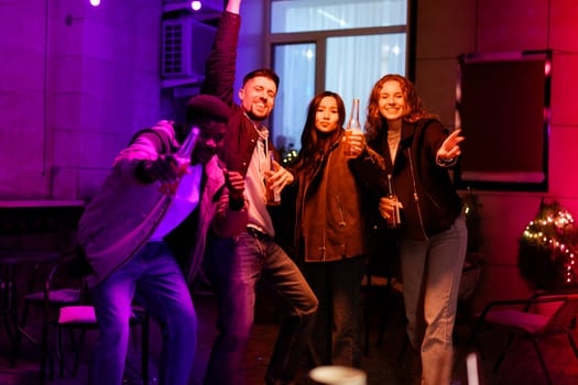 Four friends dance and celebrate together in a lively atmosphere filled with colorful lights. They enjoy drinks while showcasing their excitement and camaraderie at night.