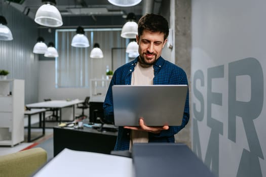 A man in a blue checkered shirt is smiling while using a laptop in a contemporary office setting. The workspace features modern decor and ample lighting.