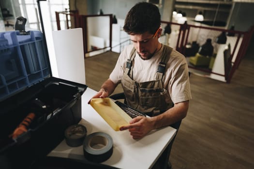 A man is focused on examining a wooden piece while seated at a workspace. Various tools and supplies are neatly arranged in a toolbox nearby, highlighting a crafting preparation in a workshop.