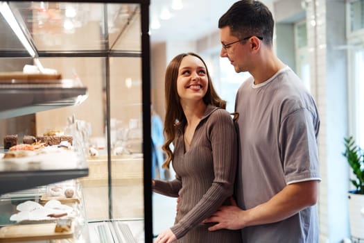 A couple stands closely in a bakery, smiling at each other while looking at a display case filled with assorted pastries and desserts. The warm atmosphere enhances their connection.