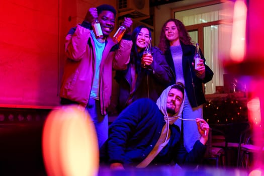 Four friends gather indoors, holding drinks while enjoying a vibrant atmosphere filled with colorful lights. They share smiles, embodying a joyous celebration together.