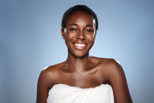 A woman with short hair smiles broadly while wearing a towel, showcasing radiant skin and confidence in a beauty treatment atmosphere with a soft blue backdrop.
