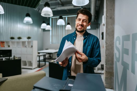 A man stands in a contemporary office environment, cheerfully sorting through papers. He wears a blue checkered shirt, surrounded by modern furnishings and warm lighting.