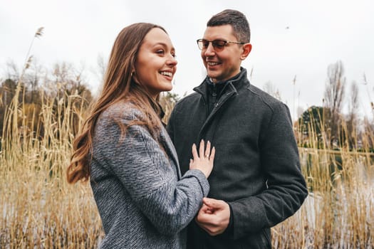A couple stands by a serene lake, smiling and holding hands. Tall grass sways gently in the breeze, adding a peaceful ambiance to their affectionate interaction on a cloudy day.