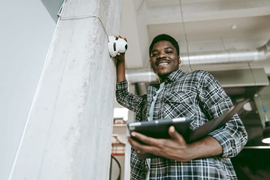 A cheerful young man stands next to a concrete pillar, holding a tablet in one hand and gesturing towards a wall light. The contemporary workspace has a vibrant atmosphere.