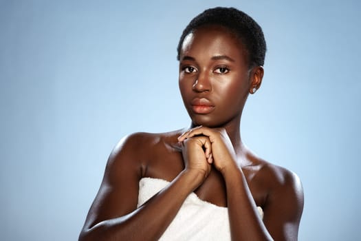 A young model stands confidently with a soft expression, wearing a white towel wrapped around her. The background is a serene shade of blue, enhancing her natural beauty and poise.