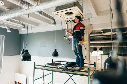 A worker is standing on a scaffold, installing an HVAC unit in a contemporary building. The space is under renovation with exposed beams and hanging light fixtures.