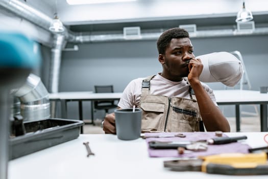 A man in work overalls sits at a table in a modern workshop. He holds a cup in one hand and looks thoughtful, surrounded by tools and equipment, during a brief pause.