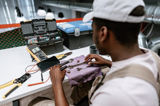 A technician focuses on maintaining various tools on a table in a workshop. He uses a multimeter and organizes wrenches while surrounded by equipment and tools.