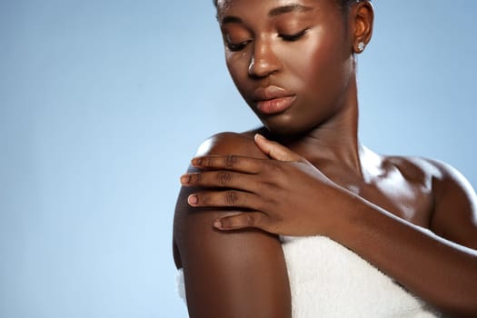 A young woman with smooth skin touches her shoulder delicately while posing. She has a serene expression and the soft light accentuates her natural beauty against a light blue backdrop.