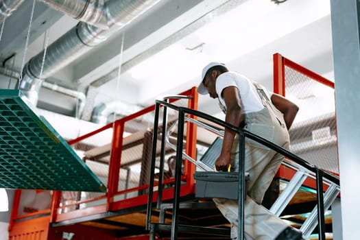 A worker is carefully climbing stairs in a well-lit industrial space, holding a toolbox. The environment features modern equipment and a busy atmosphere.