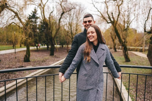 A couple happily holds hands on a bridge while smiling at each other, enjoying a serene day in a park with bare trees and a gentle stream flowing below.