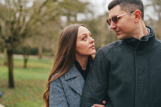 A couple stands close together in a lush park, sharing a tender moment. The woman gazes up at the man while he looks back warmly. Sunlight filters through the trees, creating a serene atmosphere.