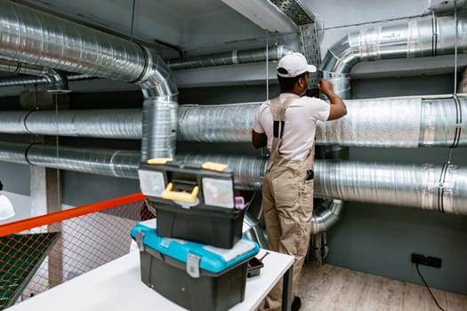 A technician works diligently on the ductwork in a spacious commercial facility. He is focused on ensuring proper installation while surrounded by metallic pipes and tools on a nearby table.