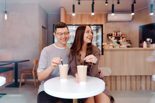 Inside a modern cafe, a couple sits at a small round table, smiling and sharing warm drinks. They are engaged in a playful moment, enjoying their relaxed afternoon together.