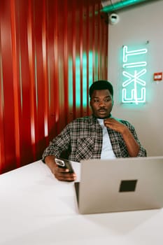 A focused young man sits at a white table, using a laptop and checking his phone in a contemporary workspace featuring red walls and bright neon signage.