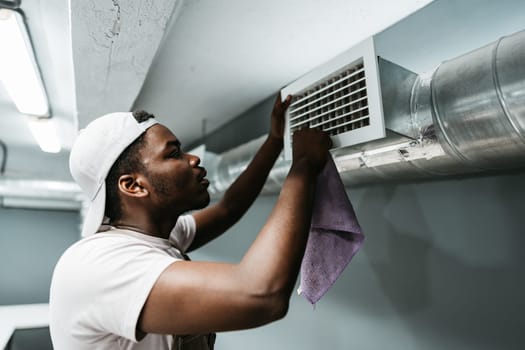 A man is focused on cleaning an air vent in a basement, using a cloth to remove dust and dirt. Proper maintenance ensures improved air quality in the enclosed space.