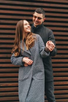 A couple smiles and embraces while standing in front of a textured wooden wall on a sunny day. The woman wears a fashionable coat, and the man is stylishly dressed.