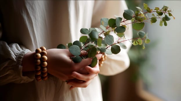 Hands holding greenery branch in peaceful indoor setting with soft lighting and wooden beads..