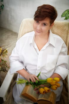 Middle-aged woman enjoys a relaxed moment with fresh tulips while reading a book in a cozy room on Women's Day.