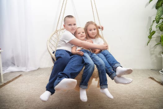 Three children sit closely together on a swing, smiling in a well-lit indoor space decorated with green plants.