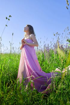 A woman is posing gracefully in a flowing pink dress amidst lush green grass and colorful flowers. The bright blue sky adds to the peaceful atmosphere of the scene.