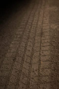 Detailed view of a tire tread imprint on a dirt road captured at golden hour. The texture of the dry soil and the diagonal pattern suggest off-road movement or agricultural activity.