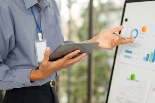 A corporate professional holds a tablet and gestures towards a digital board, showcasing data insights and analysis.