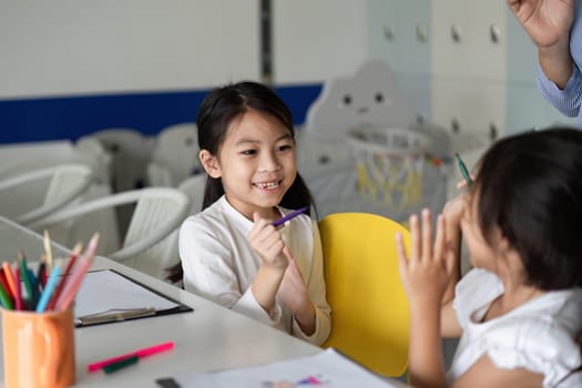 Two young girls smile and interact while sharing art supplies, fostering friendship and creativity in a classroom setting.