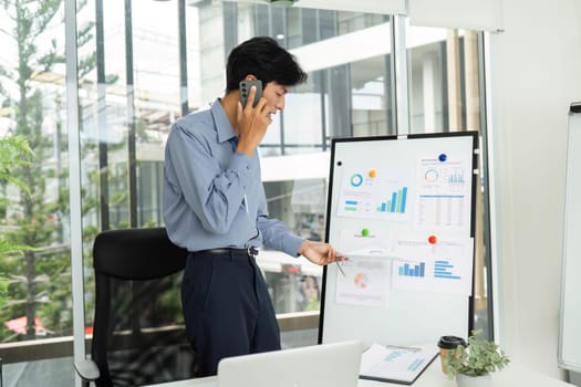 A young man presenting financial data on a board while talking on the phone, exemplifying effective multitasking in business.