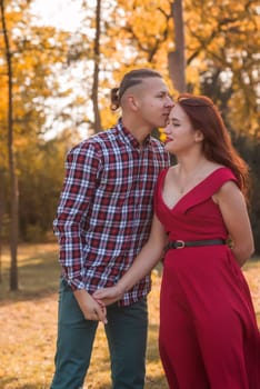Portrait of a young couple in love kissing and hugging in an autumn park. Girl in a red dress against the backdrop of golden autumn. Concept of romance, falling in love