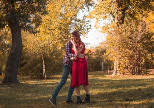 young couple in love actively relaxing in autumn park. they are running, having fun and laughing, holding hands. girl in red dress, guy in checkered shirt against golden autumn background. concept of romance