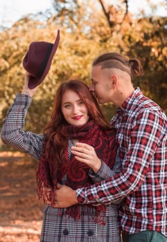 young couple in love hugging and kissing in autumn park. girl in coat, red scarf and hat in hands, against autumn trees background. concept of falling in love, relaxation