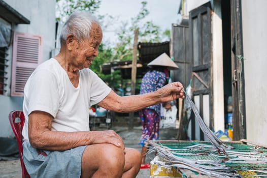Old elderly Asian Vietnamese male fisherman dries salted fish in sun in Murals Fishing Village Tam Thanh. Tam Thanh, Quang Nam province, Vietnam - April 10, 2025