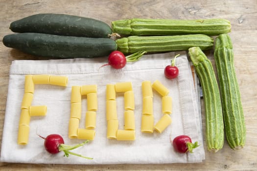 "DIET" spelled with rigatoni pasta on a white linen napkin, surrounded by fresh green vegetables (cucumbers, zucchini) and red radishes, all on a rustic wooden background. Illustrates healthy eating and balanced meals