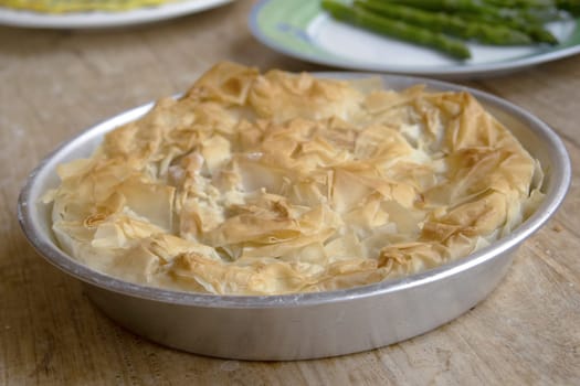 Close-up: freshly baked, golden-brown filo pastry pie in a round aluminum pan on a rustic wooden table. Highlights crispy, flaky layers. Softly blurred side dishes in background.