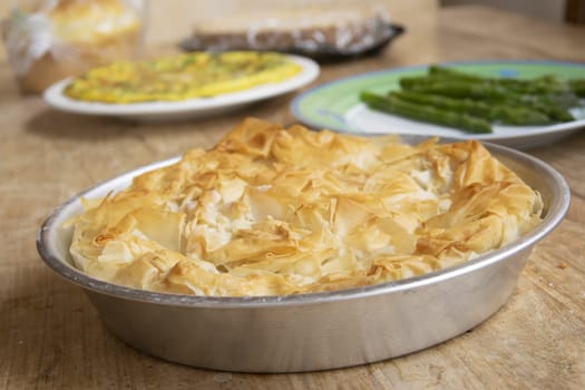 Freshly baked, golden filo pastry pie in an aluminum pan, flaky & crispy. Soft-focus background: omelette & blanched asparagus. Rustic meal setting