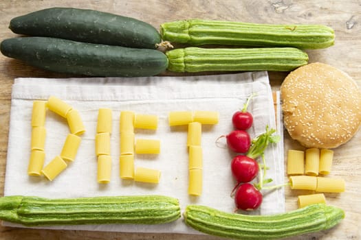"DIET" spelled with rigatoni pasta on a white linen napkin, surrounded by fresh green vegetables (cucumbers, zucchini) and red radishes, all on a rustic wooden background. Illustrates healthy eating and balanced meals