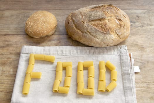 Rustic bread, sesame bun, and "FOOD" spelled with rigatoni pasta on linen, all on wood. Perfect for essential ingredients & culinary concepts