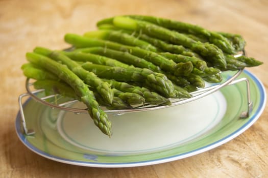 A bunch of freshly steamed asparagus spears rests on a metal rack over a green and white plate, highlighting their vibrant color and freshness