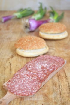 Thin slices of dry-cured salami on a wooden board with fresh bread and green onions.