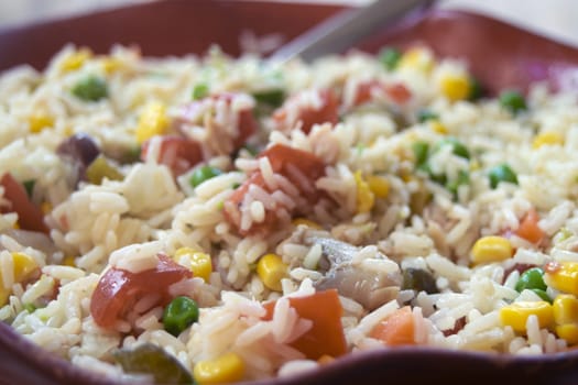 Close-up of a vibrant rice salad in a red bowl, featuring rice mixed with colorful vegetables like tomatoes, corn, peas, and possibly others. A spoon rests inside the bowl.

