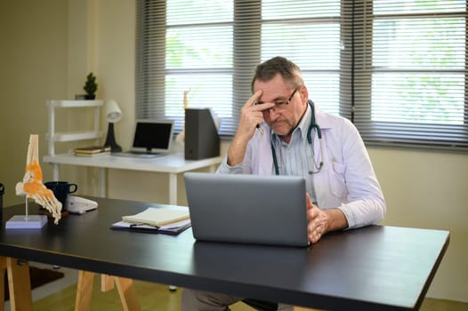 Mature doctor with stethoscope appearing exhausted during working on laptop.