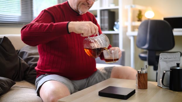 Older man in a red sweater pouring coffee from a glass pot into a cup relaxing at home.