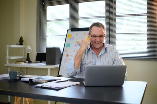 Happy mature man raising his fist in victory and looking at a laptop screen.